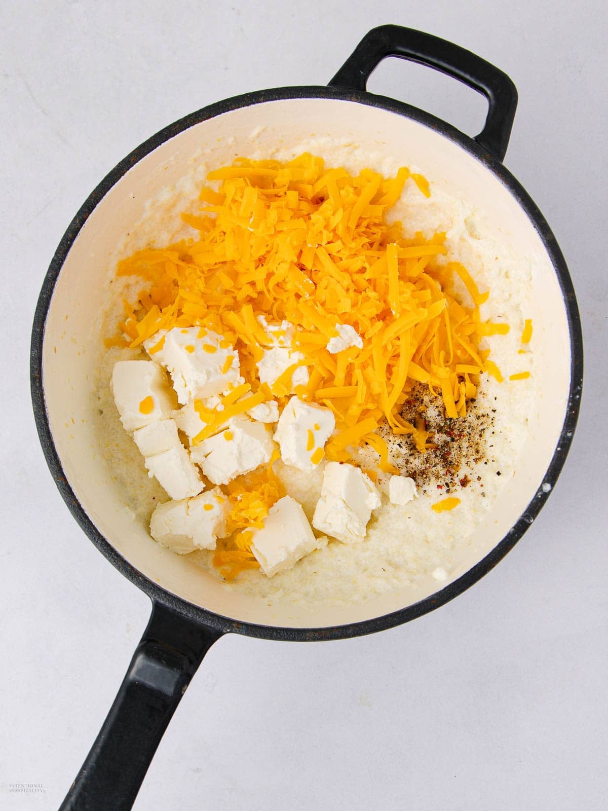 A pot contains shredded cheddar cheese, cream cheese cubes, and a creamy sauce with black pepper and red pepper flakes, ready to be mixed.