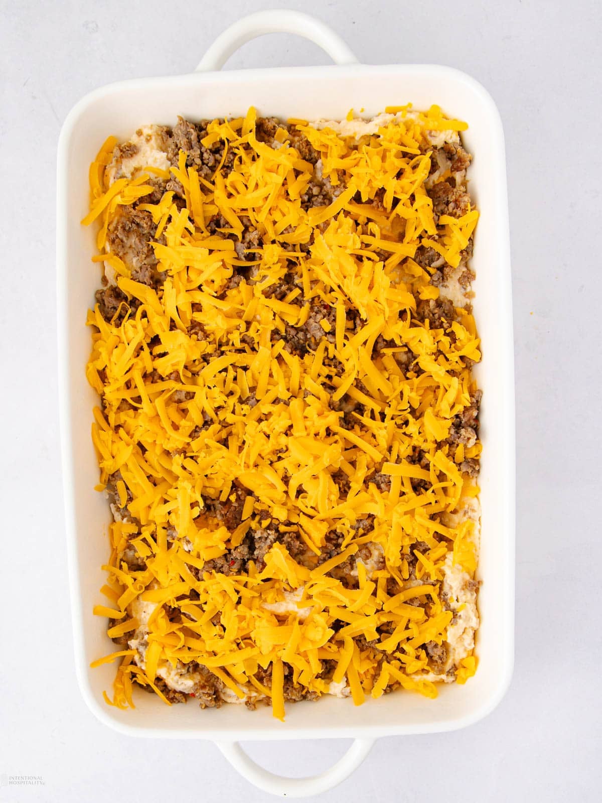 A white baking dish filled with a casserole topped with cooked ground meat and a generous layer of shredded cheddar cheese.