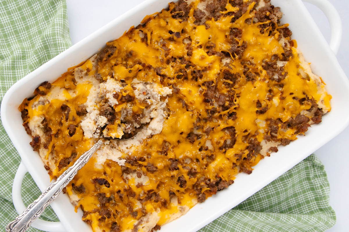 A white casserole dish filled with a cheesy ground meat and biscuit mixture sits on a green checkered cloth, with a serving spoon partially scooping out a portion.