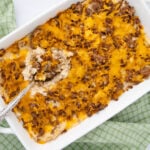 A casserole dish filled with a cheesy ground beef and potato bake, with a serving spoon scooping out a portion. The dish sits on a green checkered cloth.