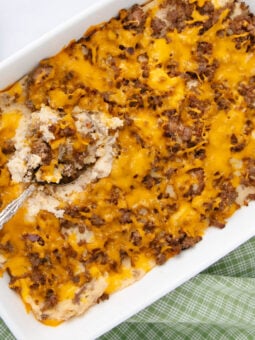 A casserole dish filled with a cheesy ground beef and potato bake, with a serving spoon scooping out a portion. The dish sits on a green checkered cloth.