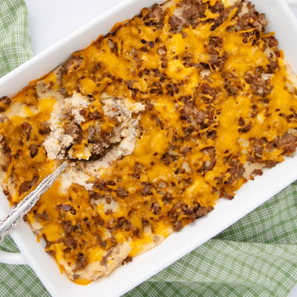 A casserole dish filled with a cheesy ground beef and potato bake, with a serving spoon scooping out a portion. The dish sits on a green checkered cloth.