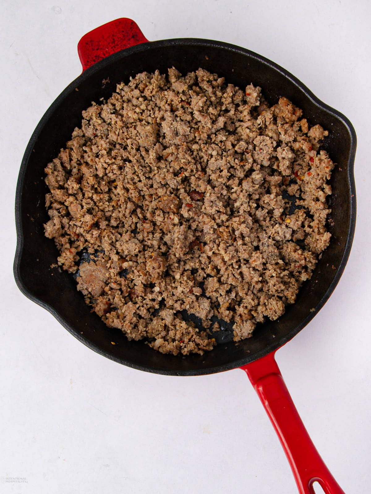 Cooked ground beef in a black cast iron skillet with a red handle, placed on a white surface.