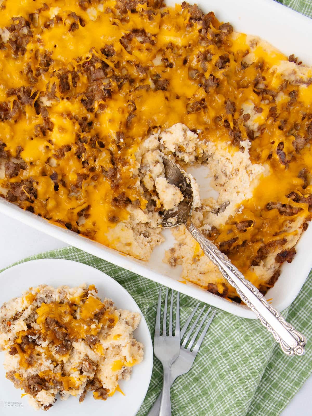 A casserole dish with a cheesy ground beef and biscuit topping, partially scooped out with a spoon. A serving sits on a plate nearby, with two forks on a green plaid cloth.