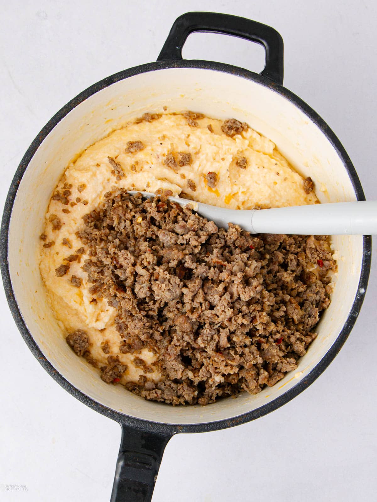 A pot of creamy yellow grits is topped with cooked ground sausage, with a white spatula mixing the ingredients together. The pot is black with a handle, sitting on a white surface.