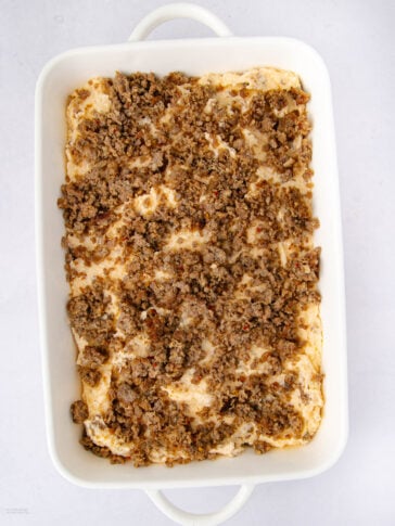 A white baking dish filled with a casserole topped with crumbled cooked sausage on a light background.