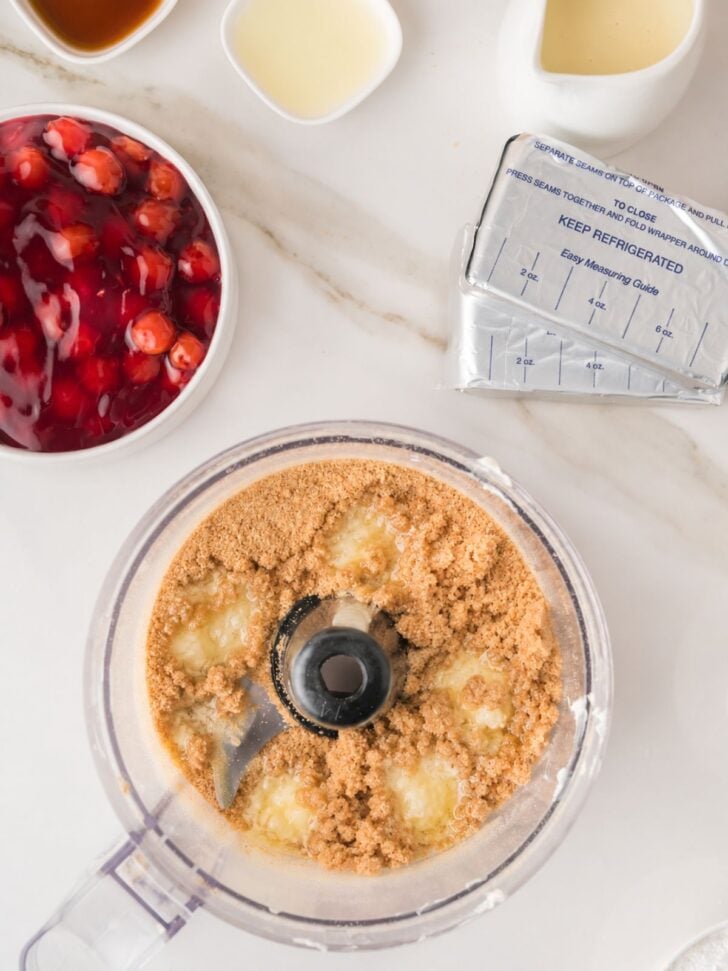 A food processor with graham cracker crumbs and butter, surrounded by a pack of cream cheese, cherry pie filling, and small bowls of other baking ingredients on a white countertop.