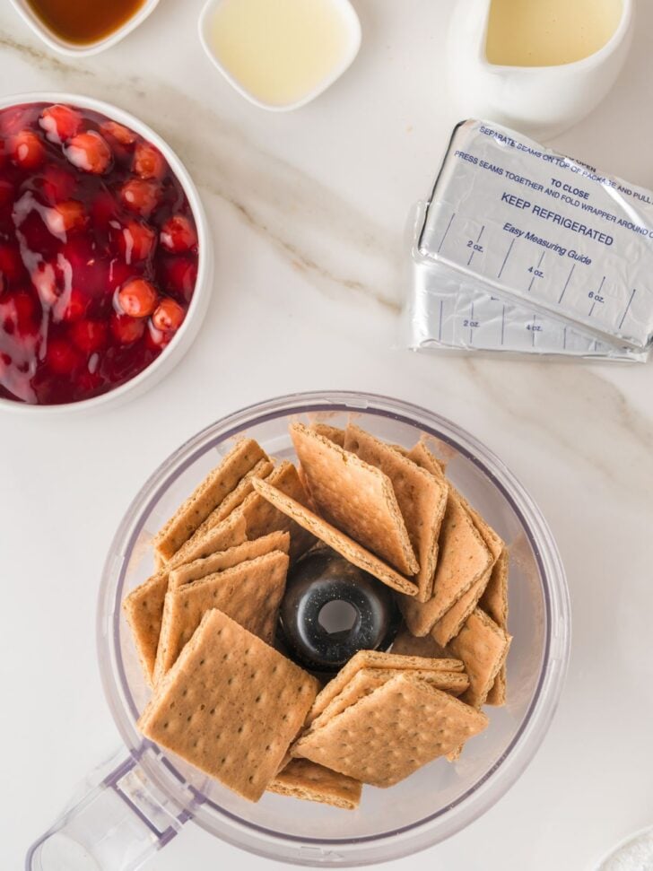 Graham crackers in a food processor bowl, with cherry pie filling, butter, sweetened condensed milk, and cream cheese packages on a white countertop.