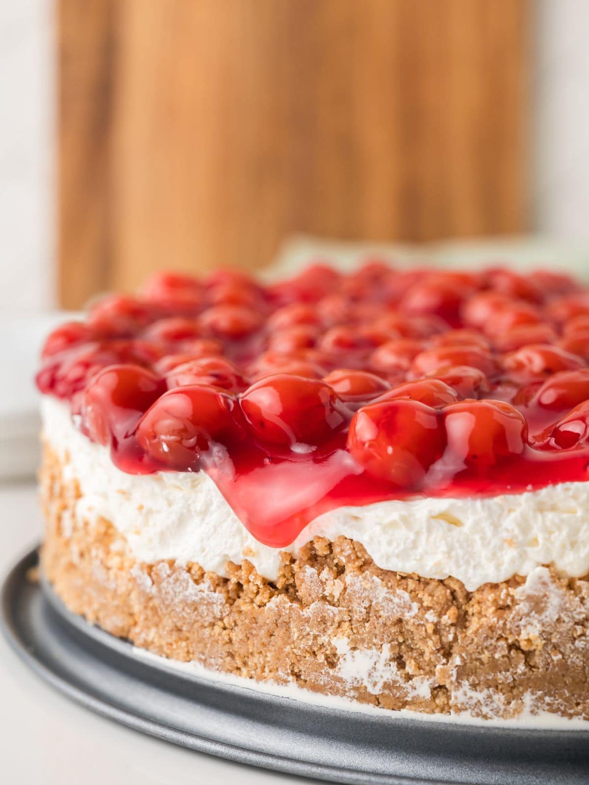 A close-up of a cherry cheesecake with a graham cracker crust, creamy filling, and bright red cherry topping, sitting on a metal platter.