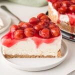 A slice of cherry cheesecake on a white plate, topped with glossy red cherries and cherry sauce, with a graham cracker crust and creamy filling.