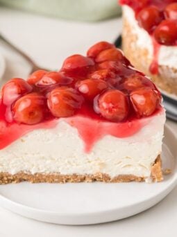 A slice of cherry cheesecake on a white plate, topped with glossy red cherries and cherry sauce, with a graham cracker crust and creamy filling.
