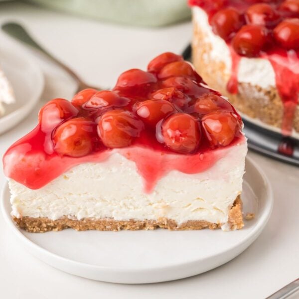 A slice of cherry cheesecake on a white plate, topped with glossy red cherries and cherry sauce, with a graham cracker crust and creamy filling.