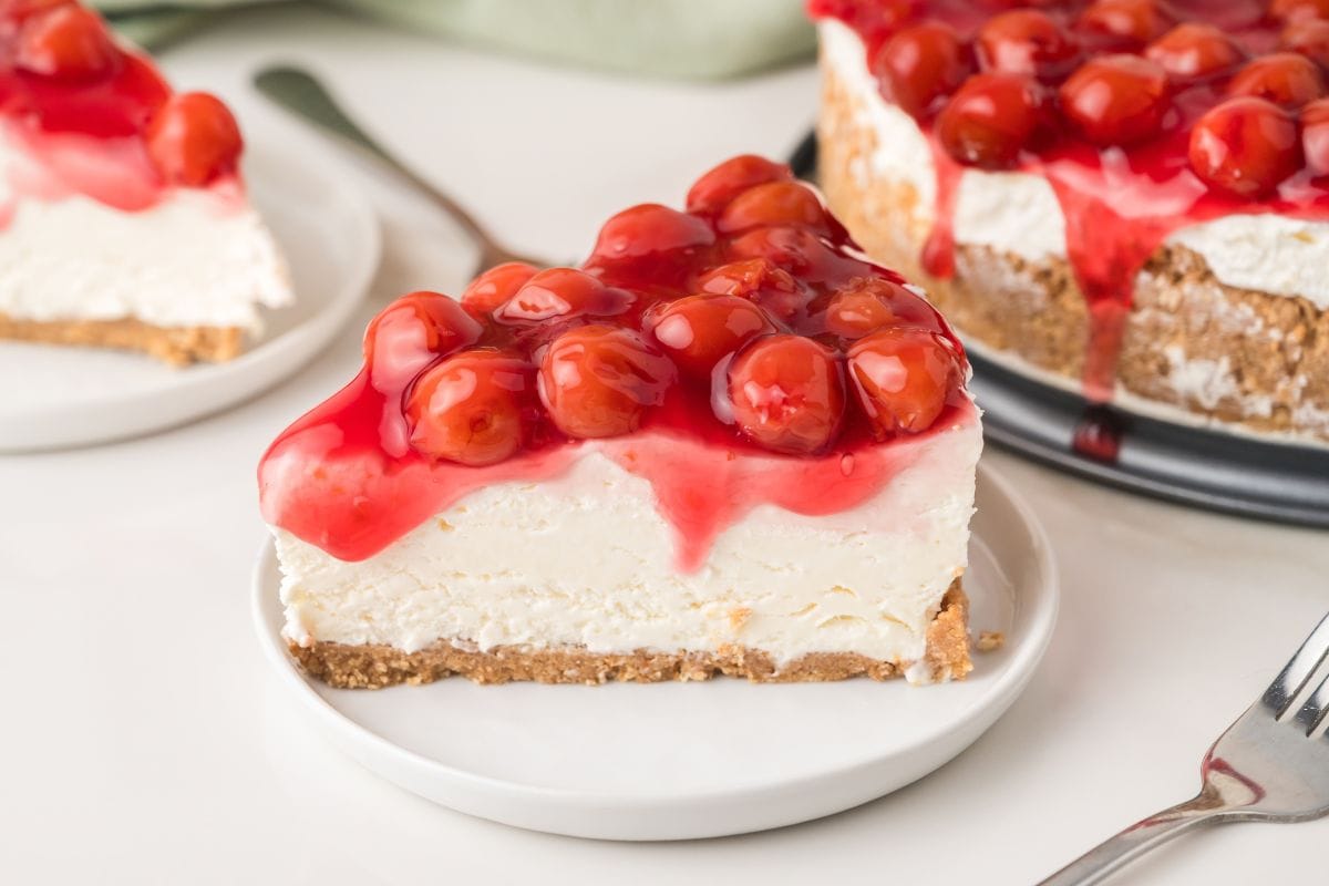 A slice of cherry cheesecake with a graham cracker crust sits on a white plate. The cheesecake is topped with whole cherries and bright red cherry glaze. The rest of the cake is in the background.