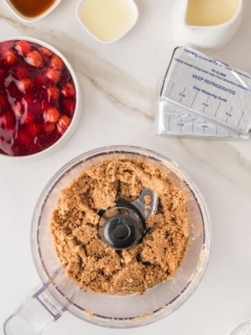 A food processor with crumb mixture sits on a white counter, surrounded by cherry pie filling, cream cheese, vanilla, and a small bowl of liquid ingredients.