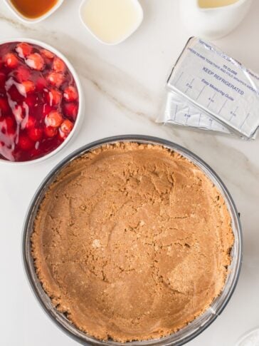 Graham cracker crust in a pan, with a packet of cream cheese, cherry pie filling, and various liquids in bowls on a white surface.