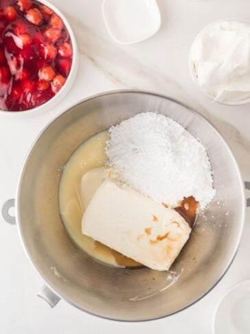 A mixing bowl with cream cheese, powdered sugar, vanilla, and condensed milk, surrounded by bowls of cherry pie filling and whipped topping on a white countertop.