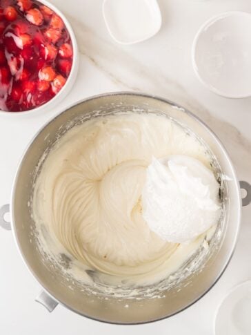 A mixing bowl with creamy batter and a dollop of whipped topping, surrounded by small white bowls and a bowl of cherry pie filling on a white marble surface.