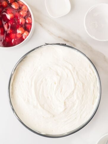 A round springform pan filled with smooth, white cheesecake batter sits on a marble surface beside a bowl of cherry topping and two empty white dishes.