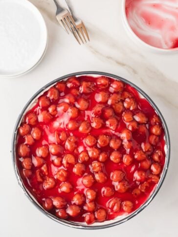 A round cherry-topped dessert, possibly cheesecake, sits on a white surface. Two forks, an empty white bowl, and a bowl with pink residue are nearby. The cherries are glossy and vibrant red.
