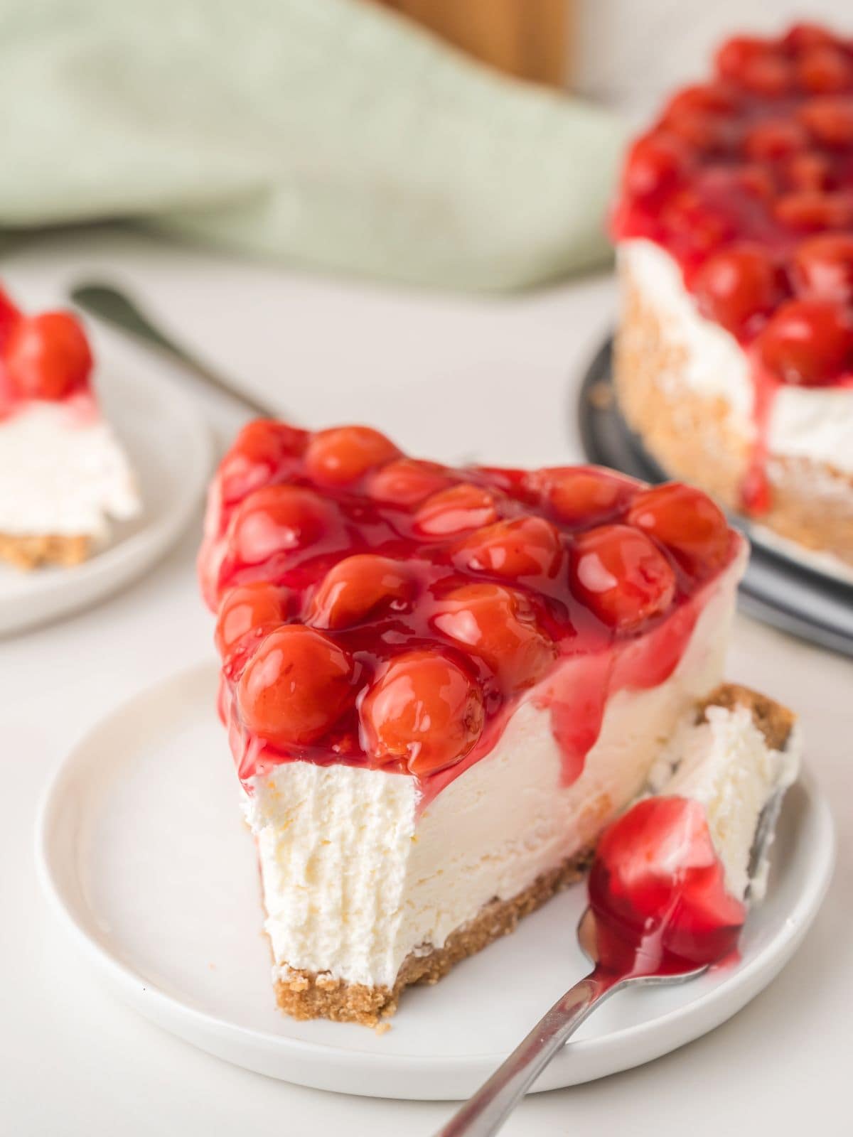 A slice of cheesecake topped with bright red cherry topping sits on a white plate, with a forkful taken out. The cheesecake has a crumbly crust and creamy filling.