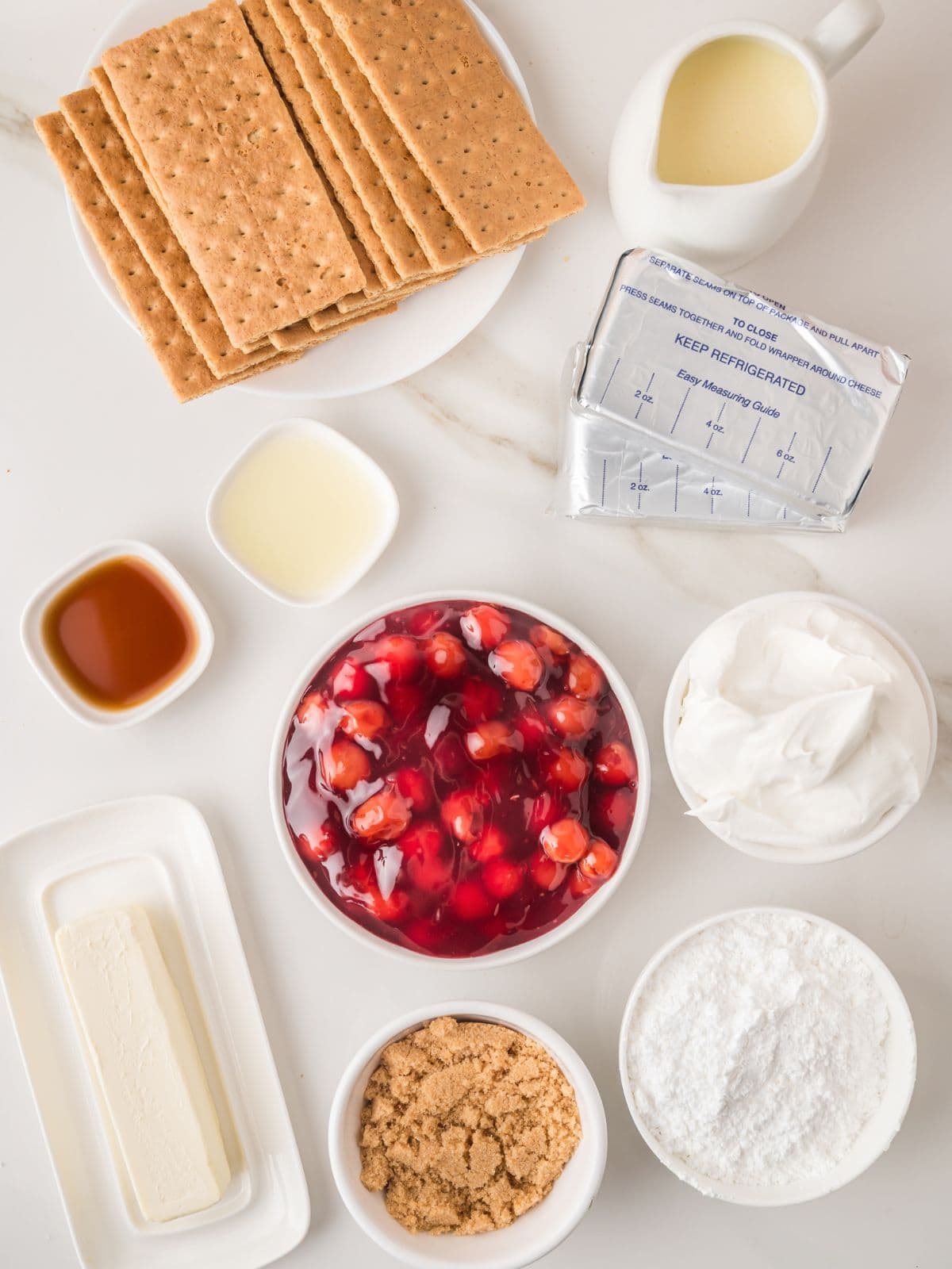 Top-down view of ingredients for a dessert, including graham crackers, cream cheese, condensed milk, vanilla extract, lemon juice, cherry pie filling, whipped topping, butter, brown sugar, and powdered sugar on a white surface.
