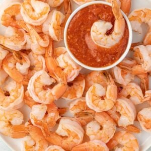 Cooked shrimp arranged around a small bowl of cocktail sauce on a white plate, with one shrimp dipped in the sauce.