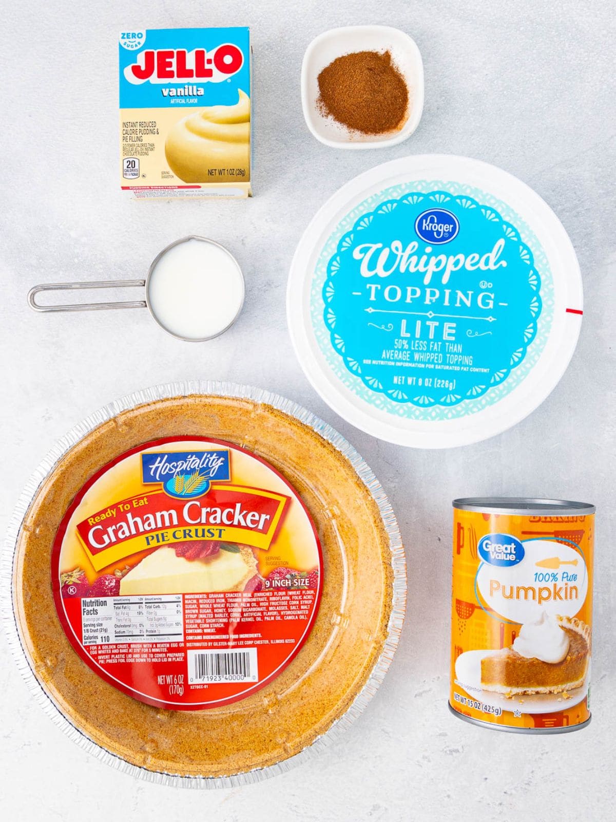 Ingredients for pumpkin pie laid out: a graham cracker pie crust, can of pumpkin, whipped topping, bowl of pumpkin pie spice, box of vanilla pudding mix, and a metal measuring cup of milk on a white surface.