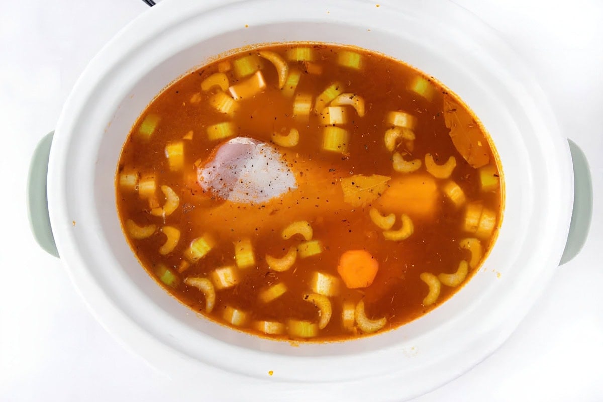 A slow cooker filled with broth, celery, carrots, a bay leaf, spices, and a raw piece of chicken, viewed from above on a white background.