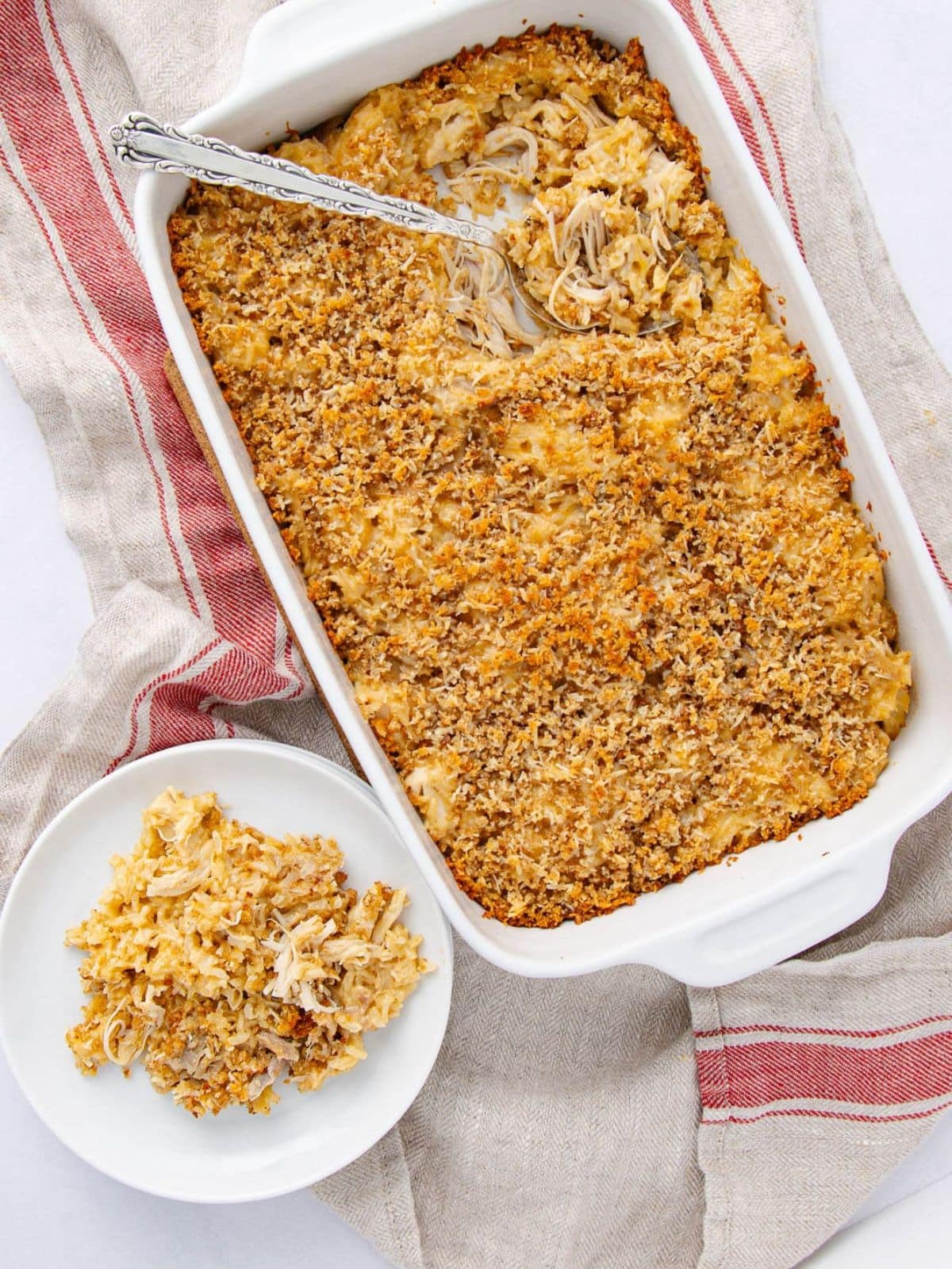 A white dish filled with baked chicken casserole topped with breadcrumbs, with a serving spoon inside. A small portion is served on a white plate beside the dish, all placed on a beige towel with red stripes.