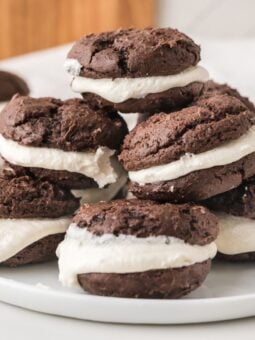 A plate of chocolate whoopie pies with white cream filling, stacked on top of each other on a white surface.