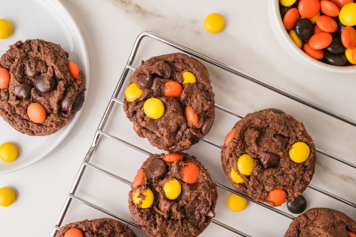 Three chocolate cookies with orange and yellow candy pieces on a wire cooling rack, with more cookies and candies on the surface nearby.