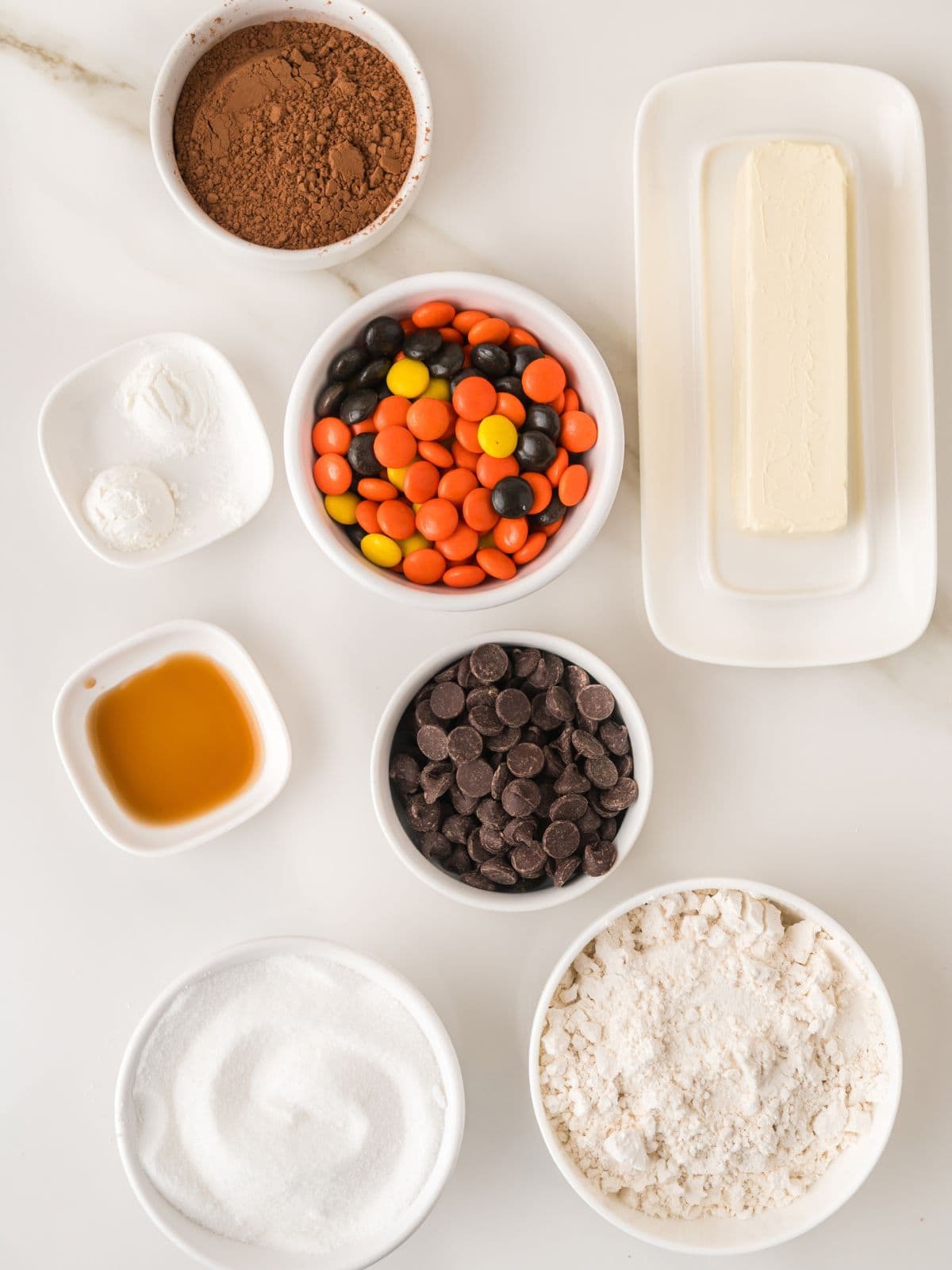 Ingredients for baking arranged in bowls and on a dish, including cocoa powder, candy-coated chocolates, a stick of butter, baking powder, vanilla extract, chocolate chips, sugar, and flour.