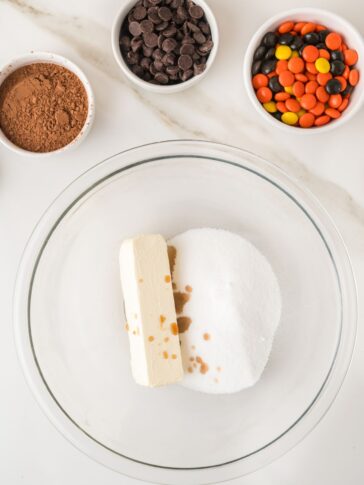 A glass bowl containing a stick of butter, sugar, and vanilla extract, with small bowls of cocoa powder, chocolate chips, and orange, yellow, and black candies nearby.