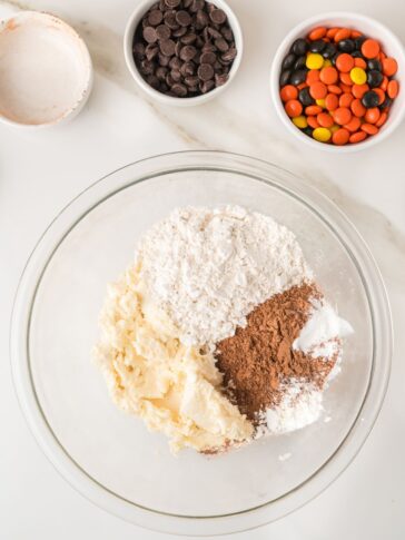 A glass bowl containing mounds of flour, cocoa powder, and creamed butter, with small bowls of chocolate chips and orange, yellow, and black candies nearby.