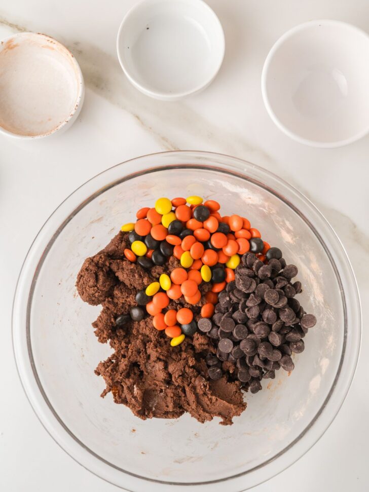 A glass mixing bowl containing chocolate cookie dough topped with a pile of orange, yellow, and black candy-coated chocolates and a pile of dark chocolate chips.