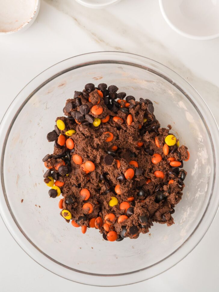 Chocolate cookie dough in a glass mixing bowl, studded with chocolate chips and orange and yellow candy-coated chocolates.