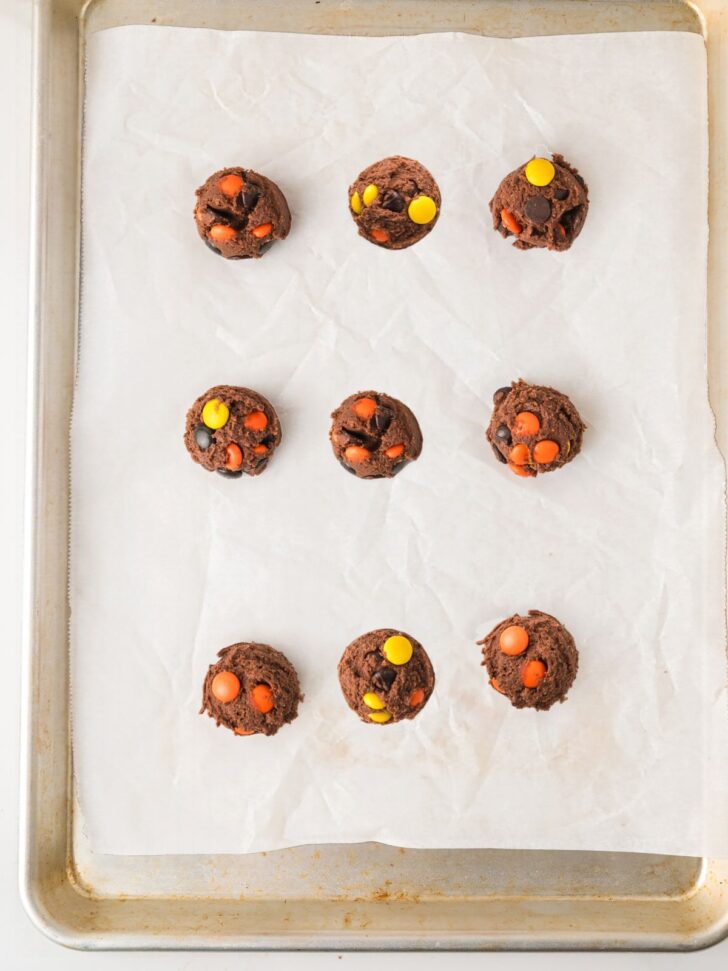Nine scoops of chocolate cookie dough studded with orange and yellow candy pieces, arranged on a parchment-lined baking sheet.