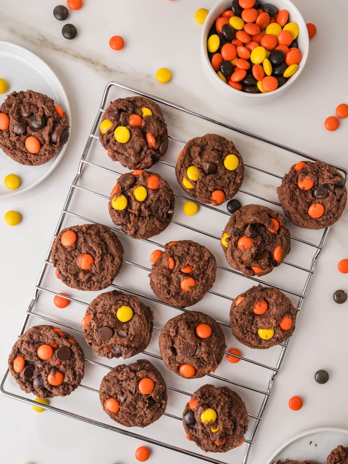 Chocolate cookies studded with orange, yellow, and black candy-coated chocolates cooling on a wire rack, with a small bowl of the candies nearby.