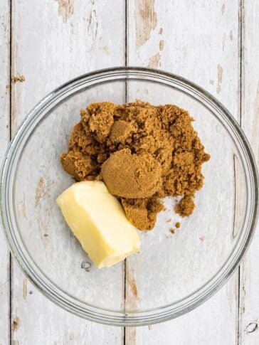 A stick of butter and a pile of brown sugar in a glass mixing bowl.