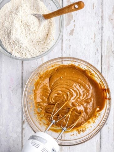 A bowl of flour with a wooden spoon and a bowl of mixed brown batter with electric beaters.
