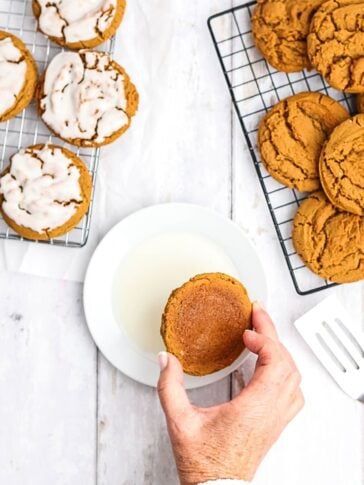 A hand dips a round cookie into a small plate of white glaze, with more glazed and plain cookies arranged on cooling racks nearby.