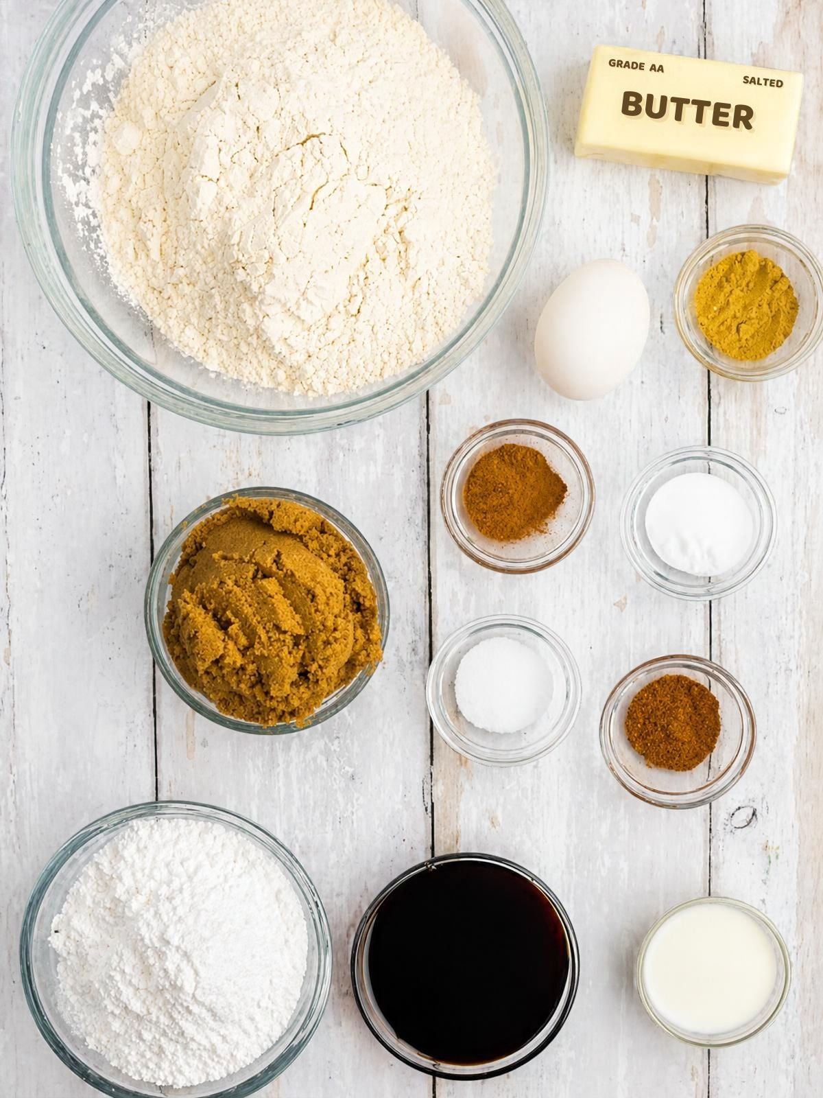 Ingredients for baking arranged on a white wooden surface, including bowls of flour, brown sugar, powdered sugar, molasses, milk, a stick of butter, an egg, and small dishes of spices and leavening agents.