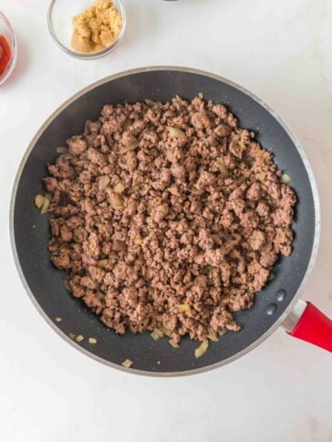 Ground meat and sautéed onions cooking in a skillet, with small bowls of brown sugar and tomato paste nearby.