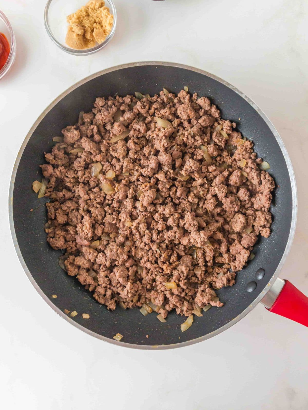Ground meat and sautéed onions cooking in a skillet, with small bowls of brown sugar and tomato paste nearby.