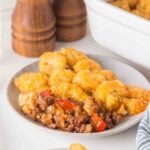 A plate of tater tot casserole with ground meat, red peppers, and melted cheese, with a baking dish and salt and pepper shakers in the background.
