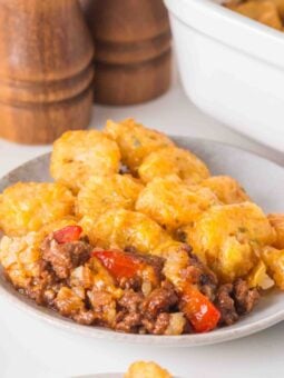 A plate of tater tot casserole with ground meat, red peppers, and melted cheese, with a baking dish and salt and pepper shakers in the background.