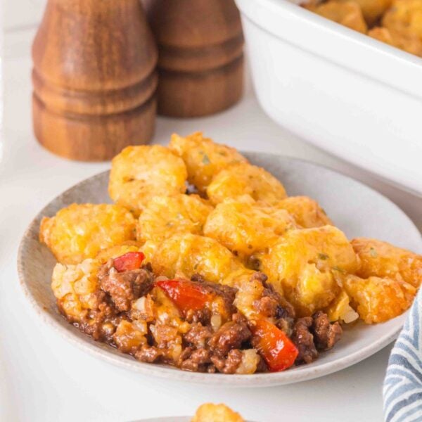 A plate of tater tot casserole with ground meat, red peppers, and melted cheese, with a baking dish and salt and pepper shakers in the background.
