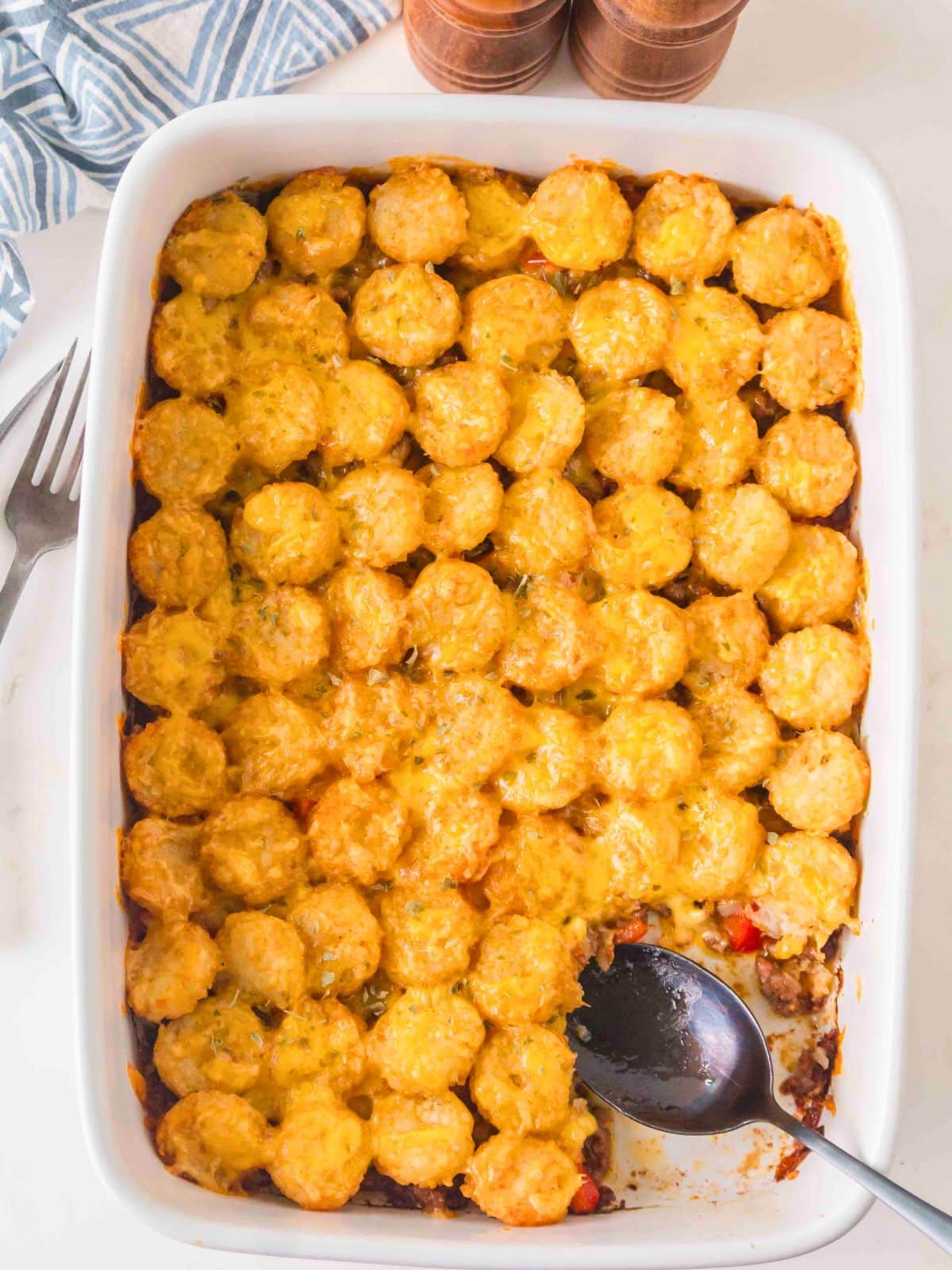 A rectangular baking dish filled with a tater tot casserole, partially scooped with a serving spoon.