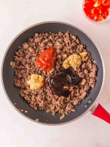 A skillet of cooked ground beef and onions topped with dollops of ketchup, mustard, brown sugar, and barbecue sauce, with a small bowl of chopped red bell peppers nearby.