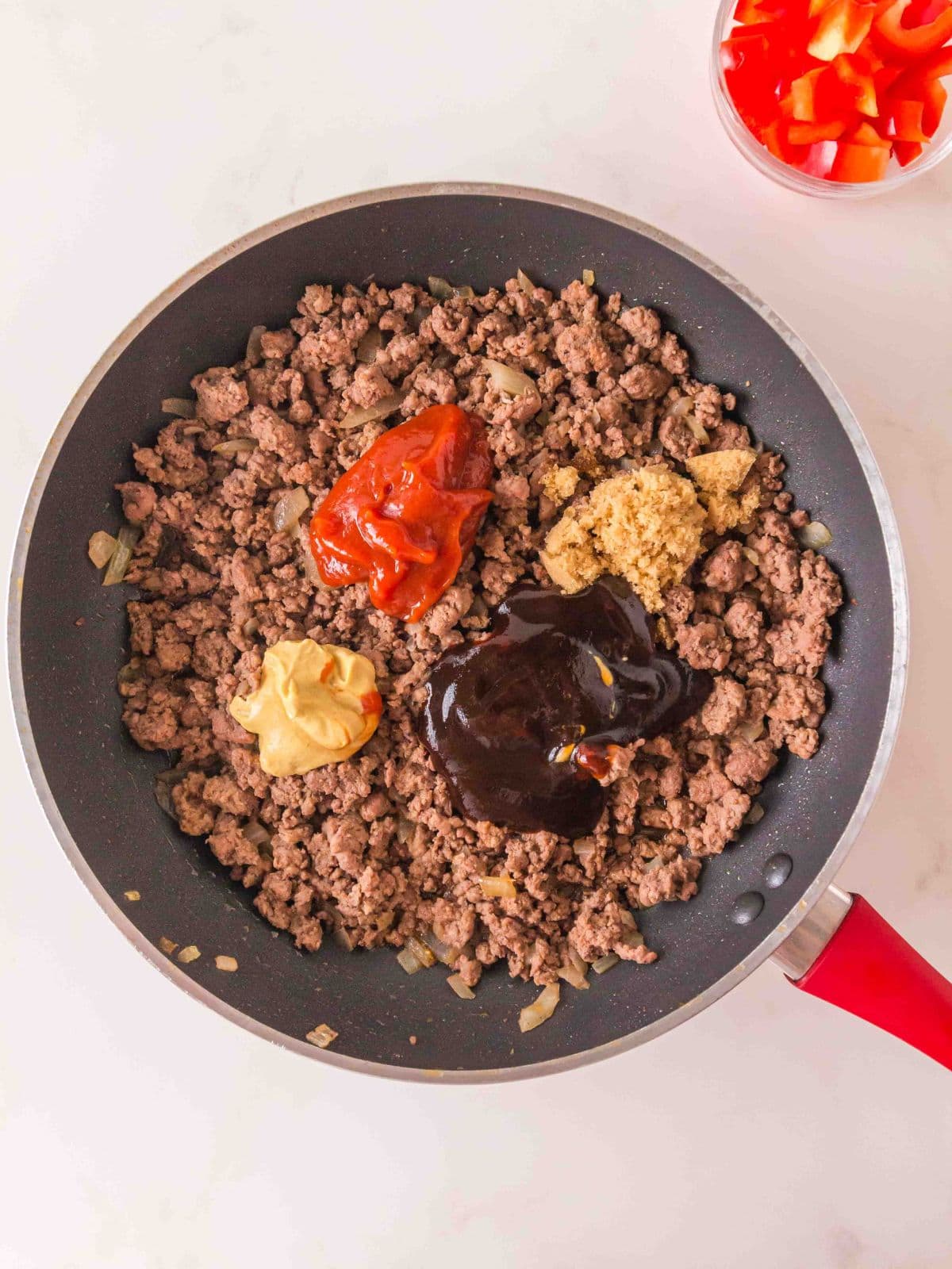 A skillet of cooked ground beef and onions topped with dollops of ketchup, mustard, brown sugar, and barbecue sauce, with a small bowl of chopped red bell peppers nearby.